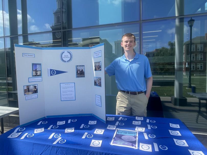 A young man in a light blue shirt stands beside a display board and table outdoors. The board features posters and photos. The mood is upbeat and promotional.